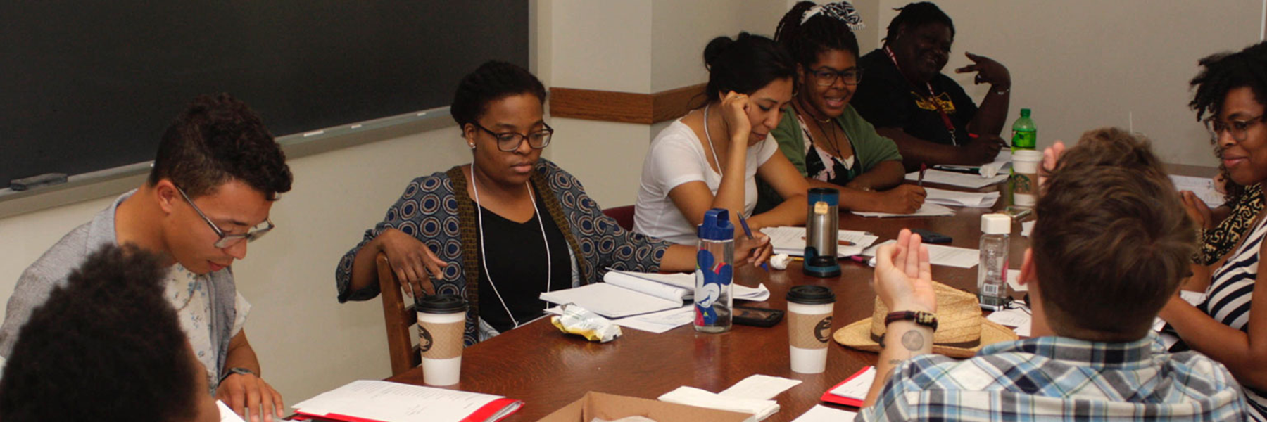 A diverse group of nine people seated around a long wooden table in a classroom. They are looking down at notes or listening to someone off-camera. Papers, notebooks, and coffee cups are scattered on the table. A chalkboard is visible in the background.