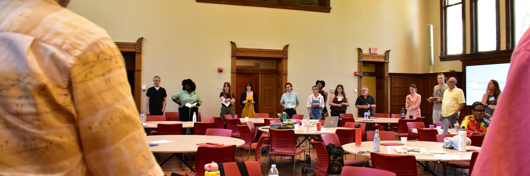 A wide-angle, eye-level shot of a large, high-ceilinged room. A group of about 15 people are standing in a semi-circle in the background. In the foreground, a person is partially visible from the back, leaning over a table with a laptop. A few other round tables are visible, set up with red chairs. Tall, arched windows and wood-trimmed doorways are visible in the background.