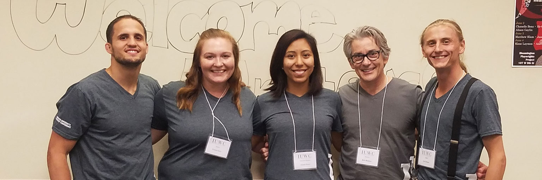 A horizontal photo of five people standing in a row and smiling at the camera. The three women and two men are wearing gray t-shirts and lanyards.